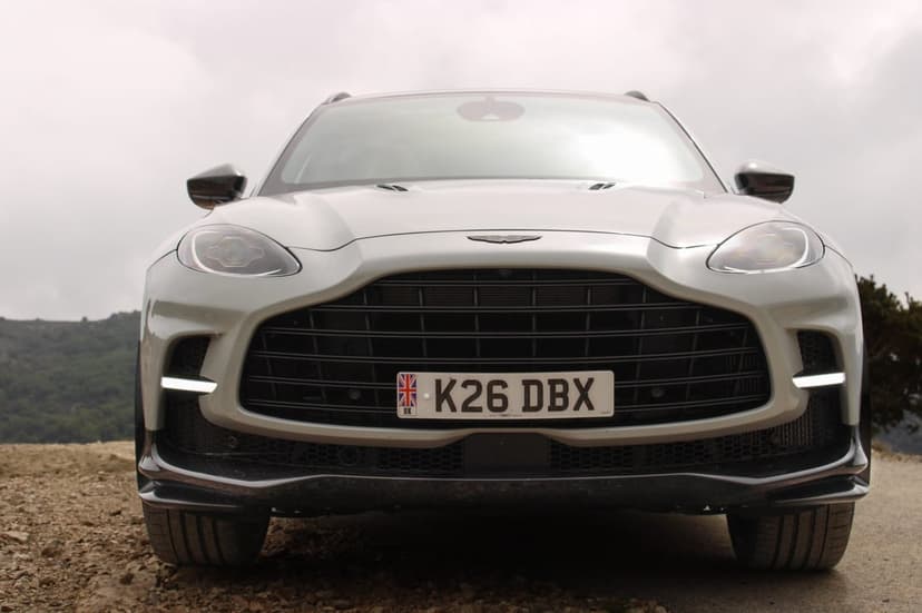 Silver Aston Martin DBX front view with wide grille and UK license plate on a gravel road under a cloudy sky.