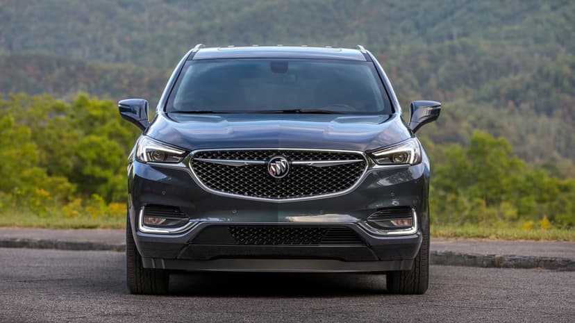 Front view of a dark grey Buick SUV parked on an asphalt road with a blurred forest mountain background.