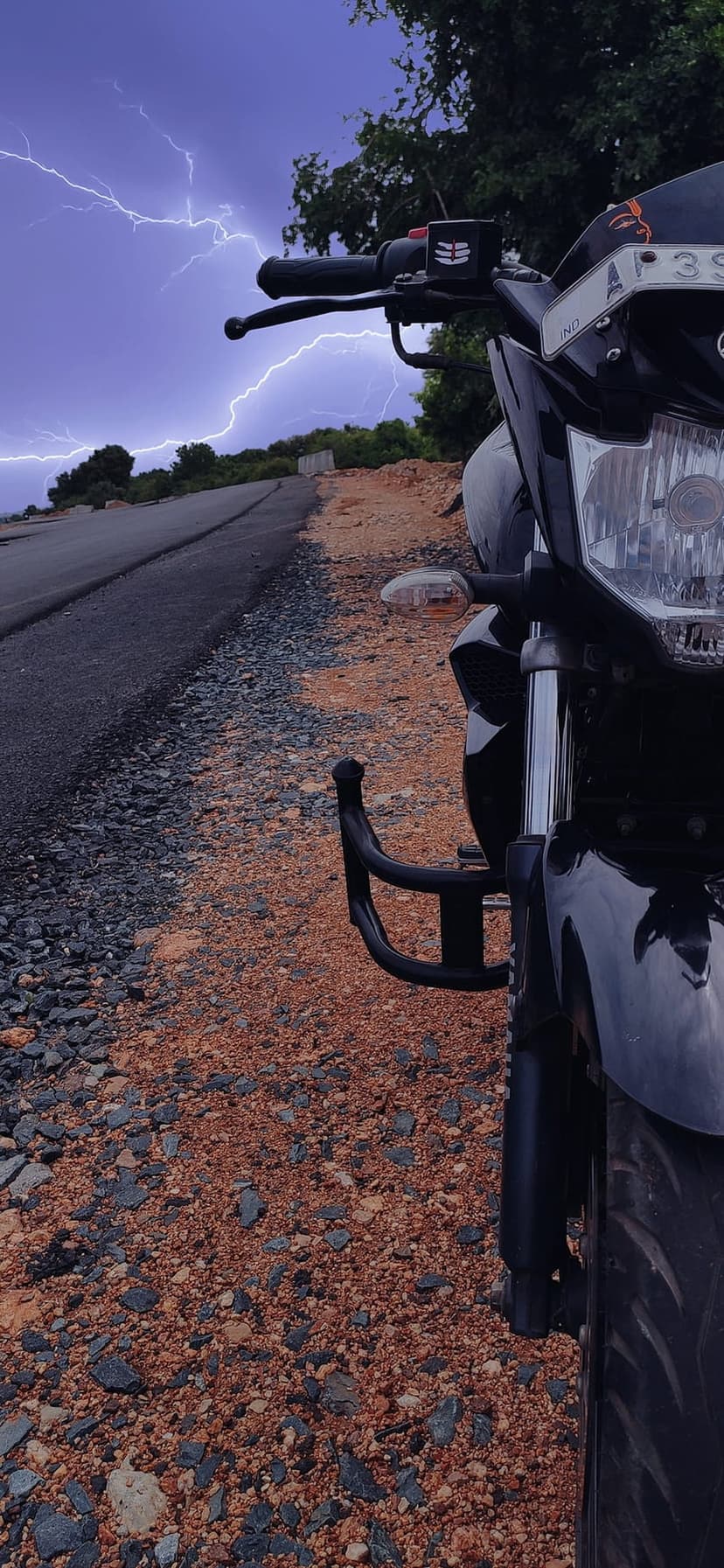 Motorcycle parked on an asphalt road while bright lightning strikes illuminate a dark, stormy night sky.