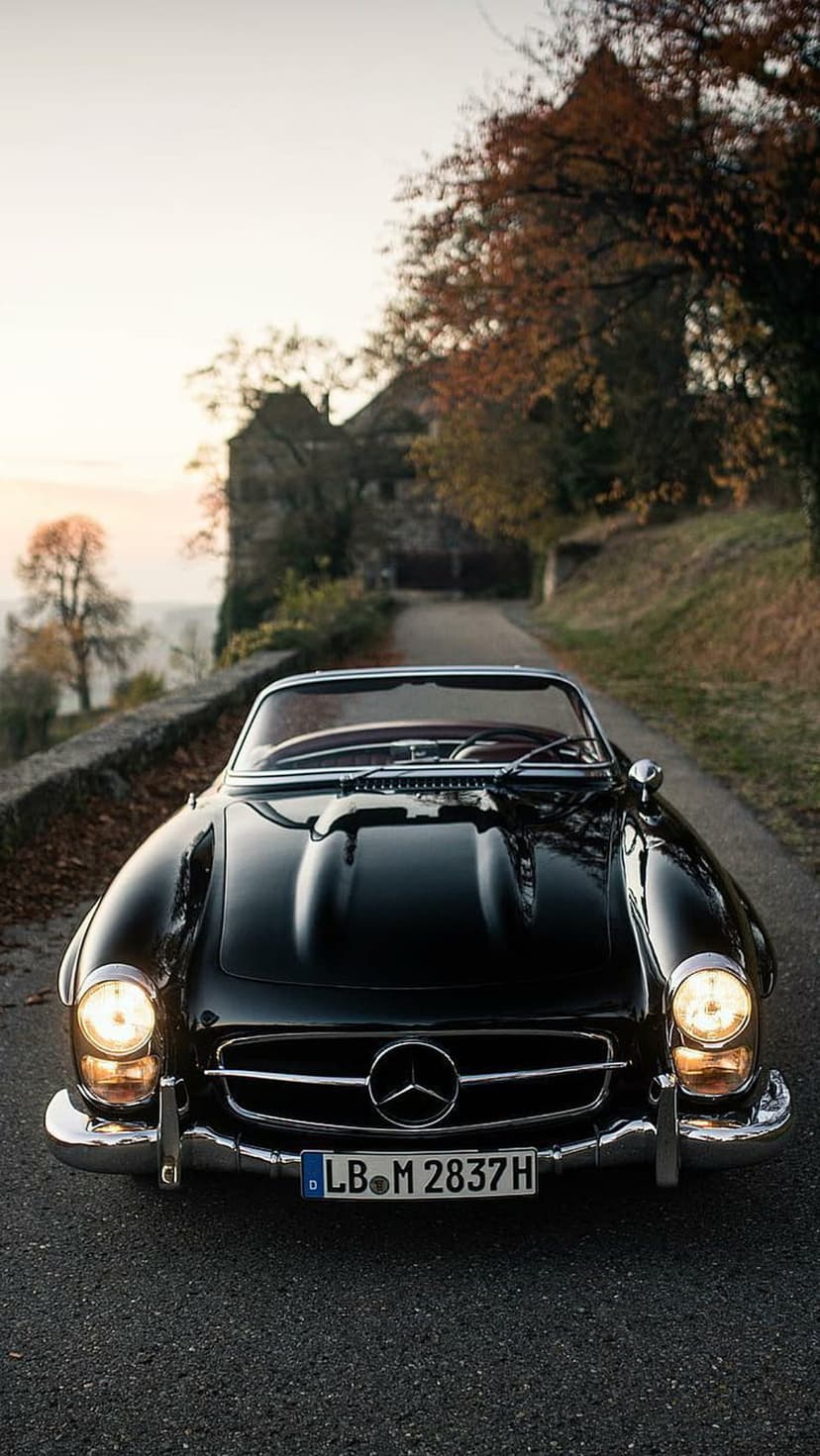 Black vintage Mercedes convertible driving on a tree-lined road with autumn leaves and a stone building behind.