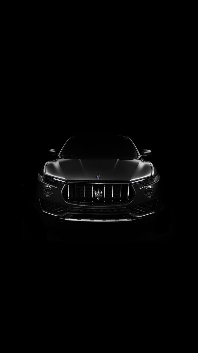 Maserati SUV front grille with glowing LED headlights and a silver trident emblem against pitch-black shadows.