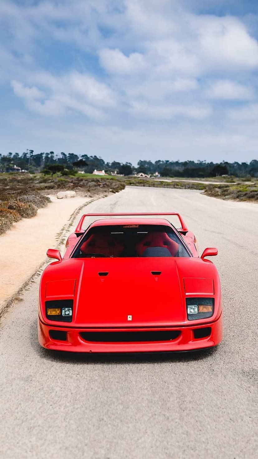 Red Ferrari F40 supercar parked on a winding road with rolling hills and a cloudy blue sky in the distance.