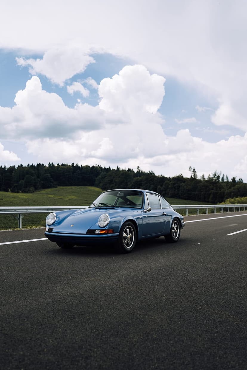 Blue vintage Porsche 911 driving on a highway through a green forest under a dramatic and moody cloudy sky.