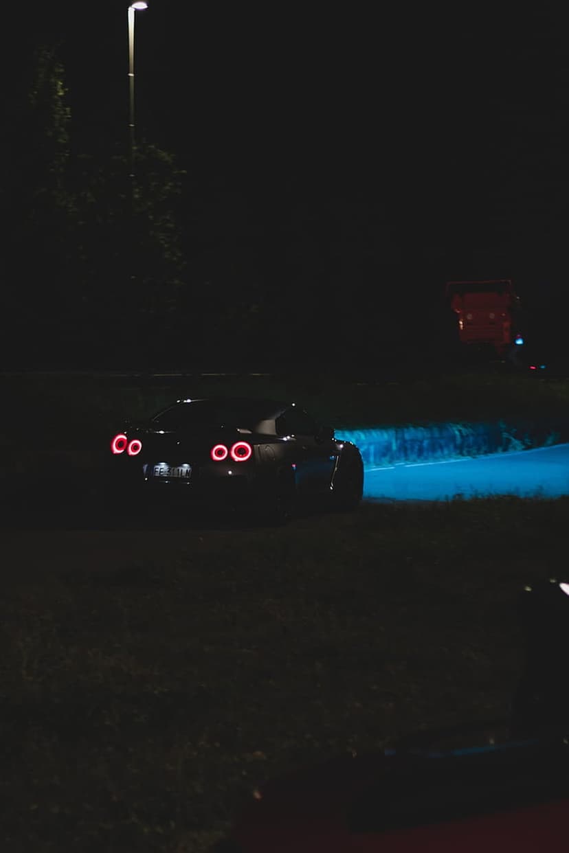 Red sports car tail lights illuminate a dark roadside with a soft crimson glow and blue accents at night.