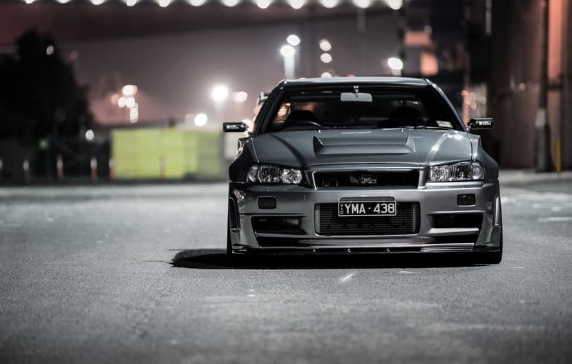 Grey Nissan Skyline GT-R R34 with a vented hood parked on a dark asphalt road with blurred city lights at night.
