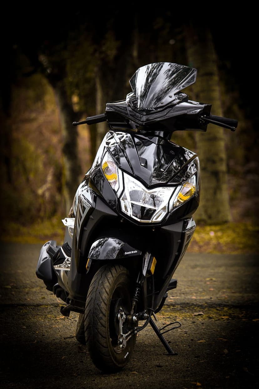 Black scooter with yellow headlight accents parked on a dirt path in a blurred forest, low-angle view.