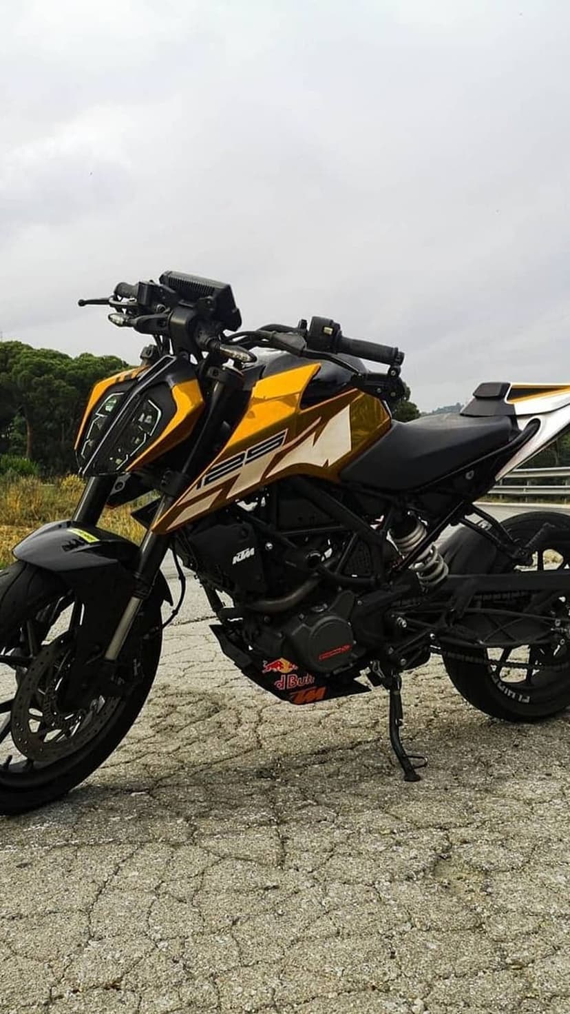 KTM Duke 125 motorcycle with gold graphics parked on a cracked road under a cloudy sky with blurred trees.