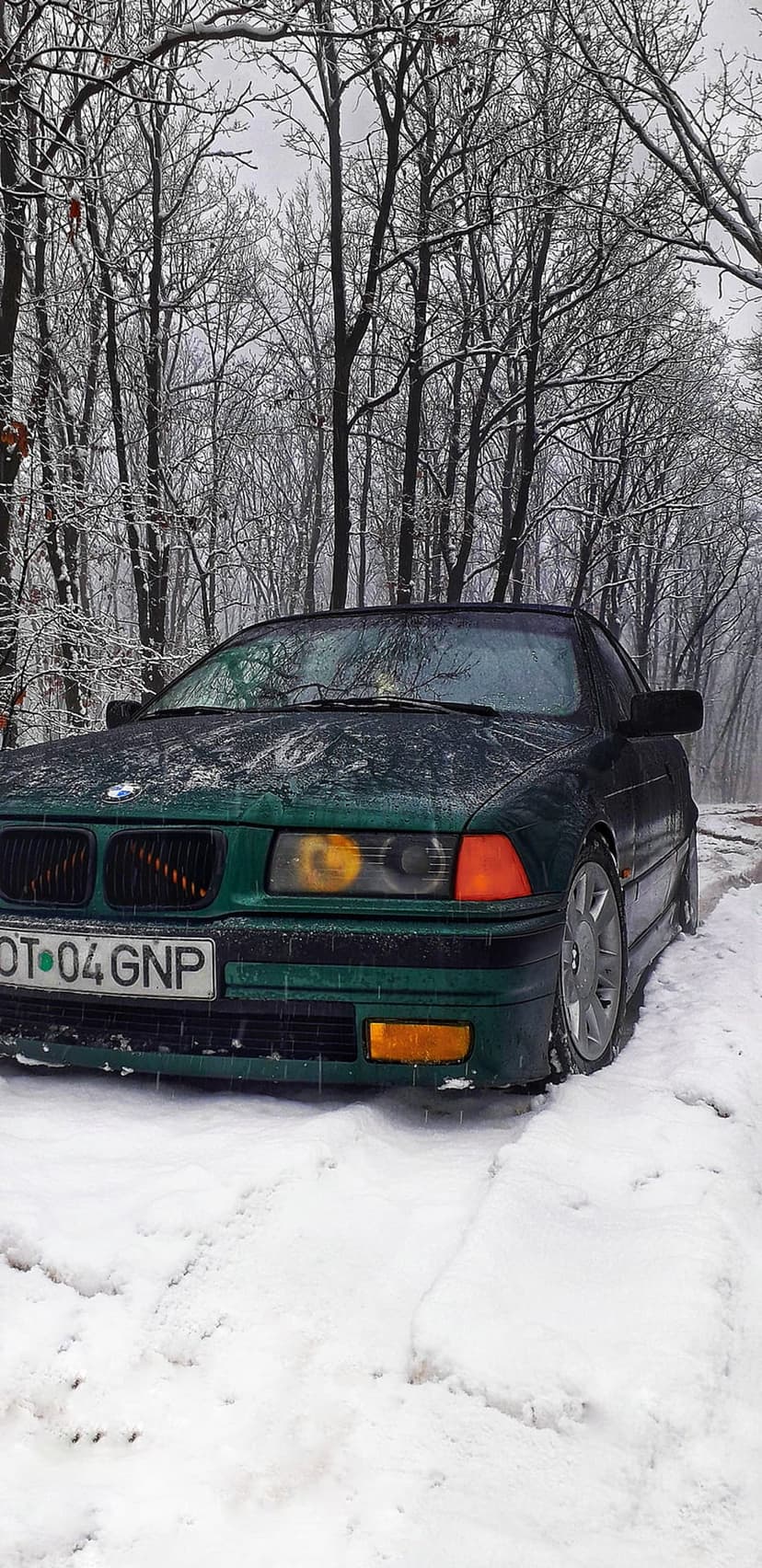 Dark green BMW E36 sedan parked on a snow-covered forest path with frost-covered trees and glowing headlights.