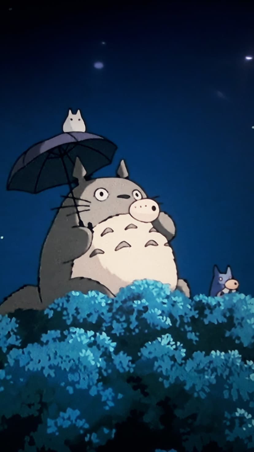 Totoro holds a wide umbrella over forest spirits amidst glowing blue bushes under a starry night sky.
