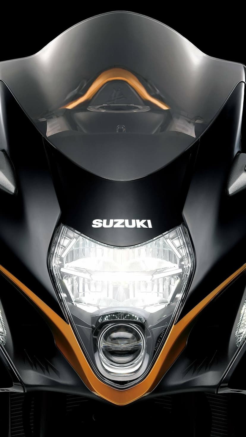 Close-up of a black Suzuki motorcycle front with a glowing LED headlight and bronze accents on a dark backdrop.
