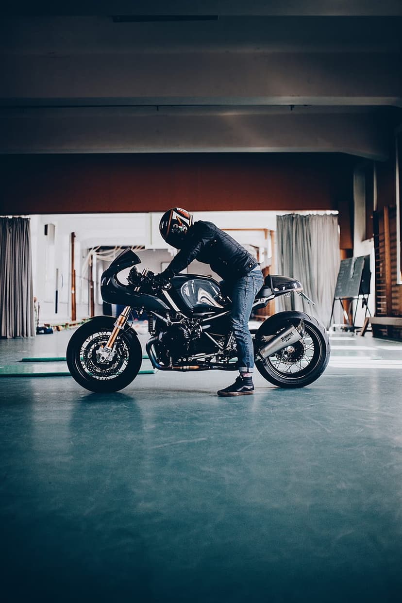 Man in a black helmet and leather jacket leans against a dark grey cafe racer motorcycle on a blue-grey floor.