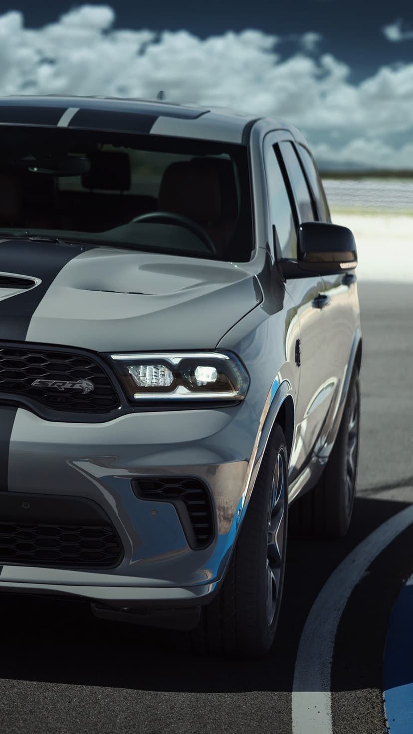 Silver Dodge Durango SRT with black racing stripes and glowing headlights against a cloudy blue sky.