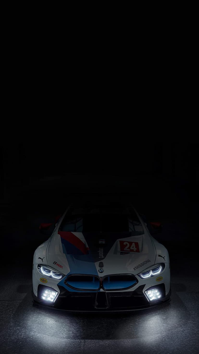 BMW M8 GTE race car with illuminated LED headlights and number 24 decals on a polished dark studio floor.