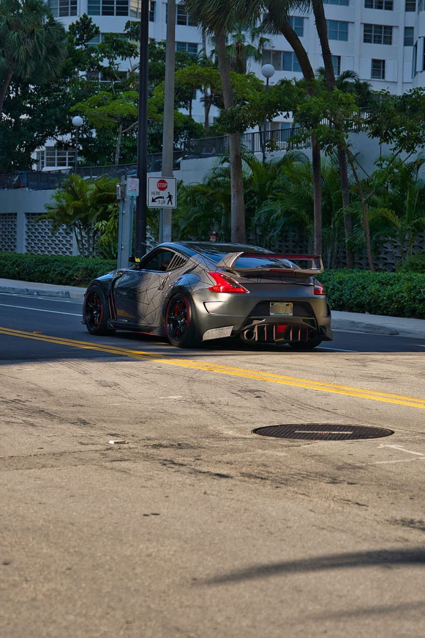 Nissan 370Z with a rear wing drives past palm trees on a sunlit city street with long shadows on the road.