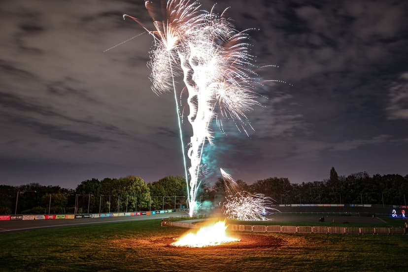 White fireworks explode in a dark sky above a bright orange bonfire on a grassy field with stadium lights behind.