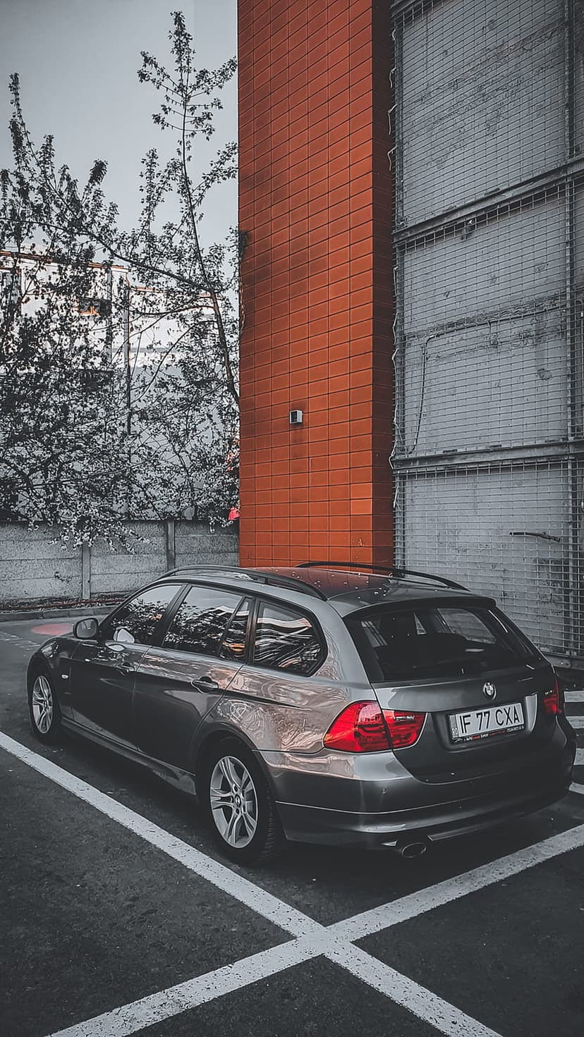 Dark gray BMW estate car parked beside a modern orange-red brick facade and industrial gray wall under bare trees.