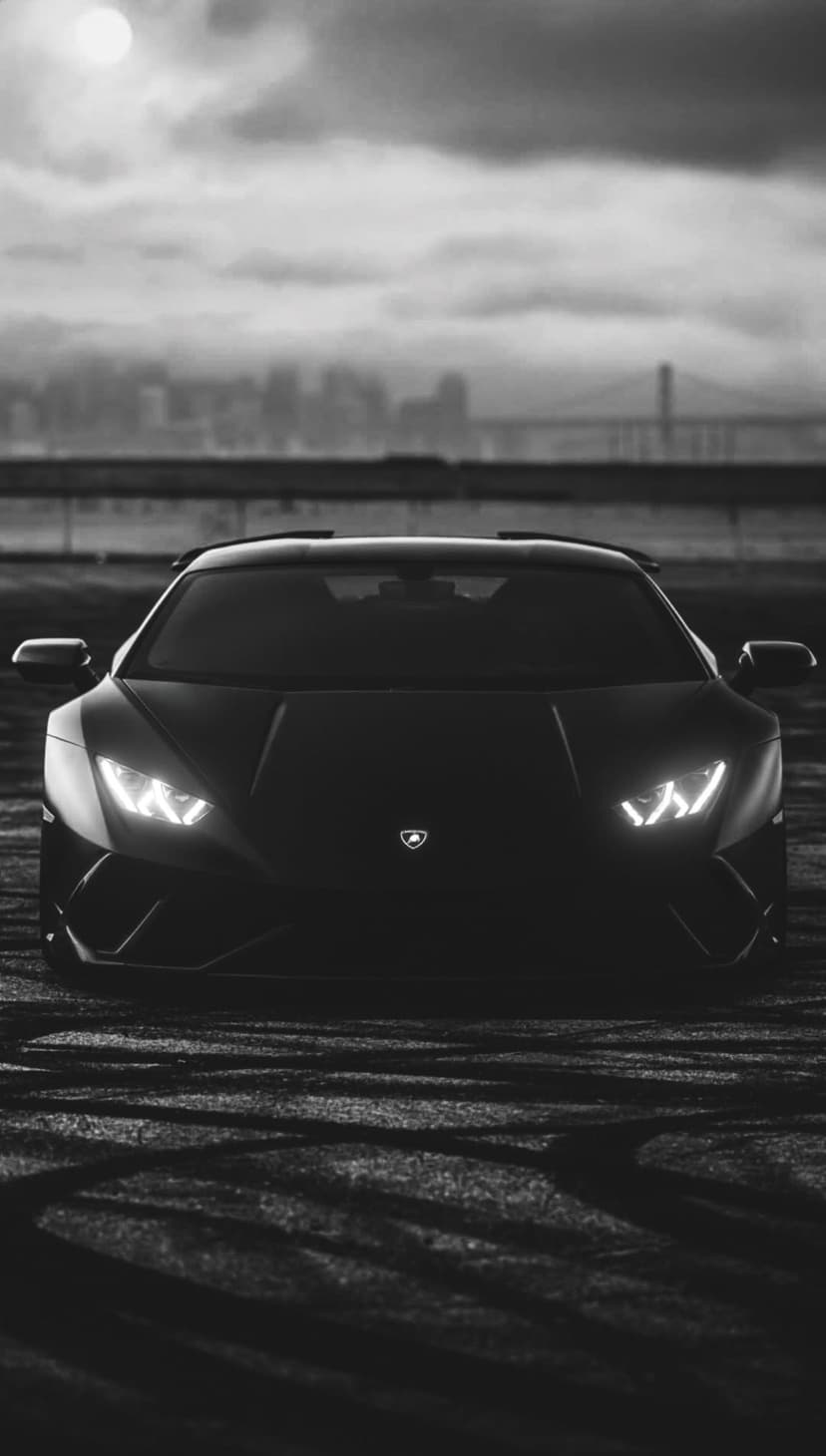 Black Lamborghini with glowing headlights on a textured road with a bridge and misty city skyline behind.