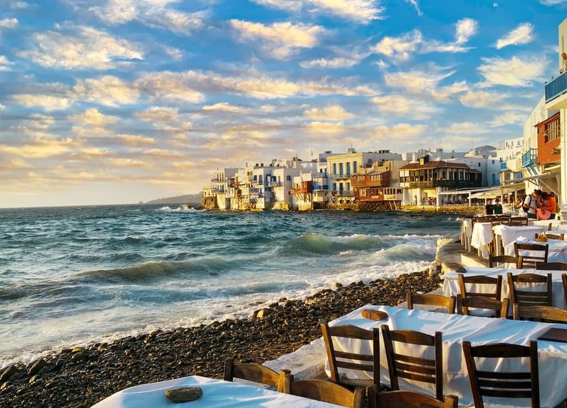 White-washed seaside restaurant tables overlook the Aegean Sea at sunset under a dramatic cloudy sky.