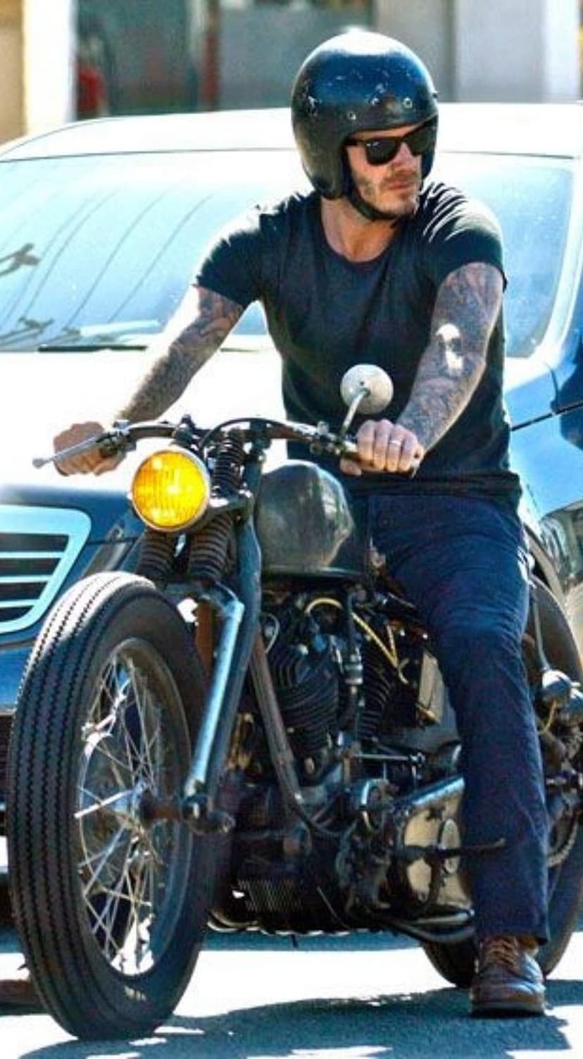 Tattooed man in a black helmet rides a vintage motorcycle with a glowing yellow headlight on a city street.