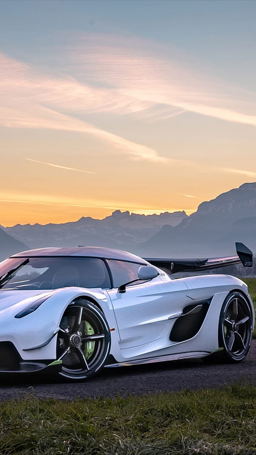 White Koenigsegg hypercar parked on a scenic road with glowing sunset mountains and orange sky in the background.