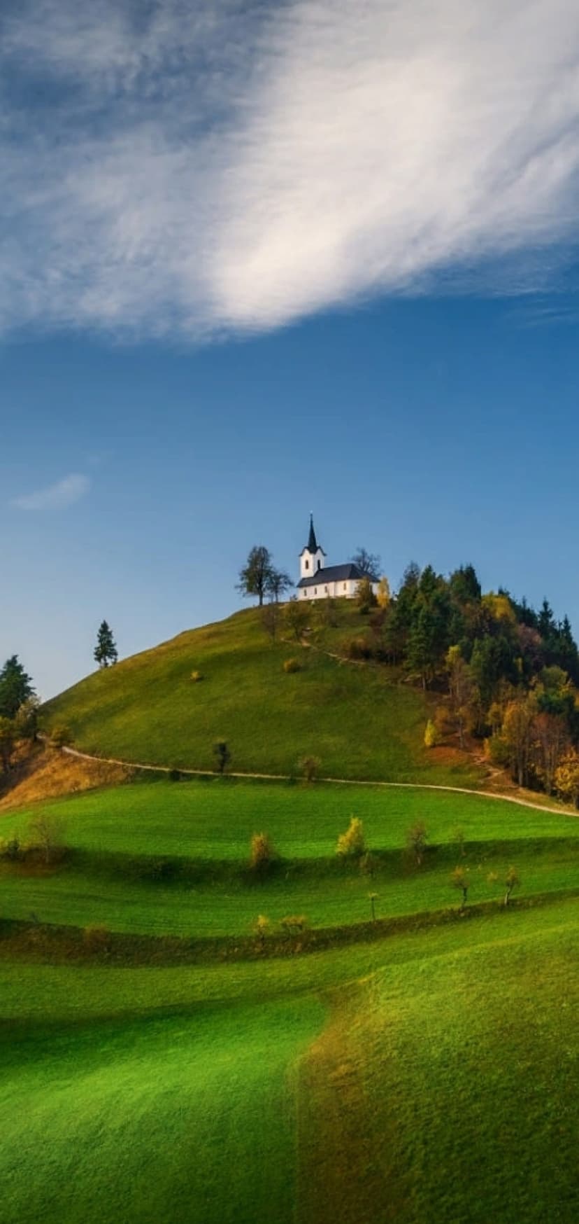 White chapel with a dark spire stands on a grassy hill under a bright blue sky with wispy clouds and autumn trees.