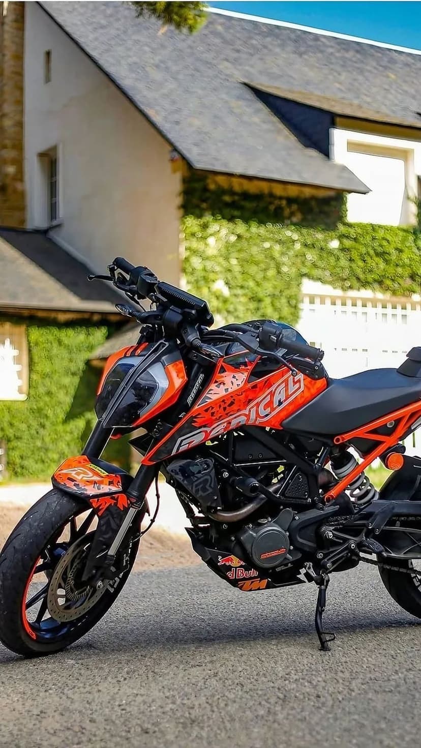 Orange KTM Duke motorcycle with radical graphics parked on asphalt before a house covered in green climbing ivy.