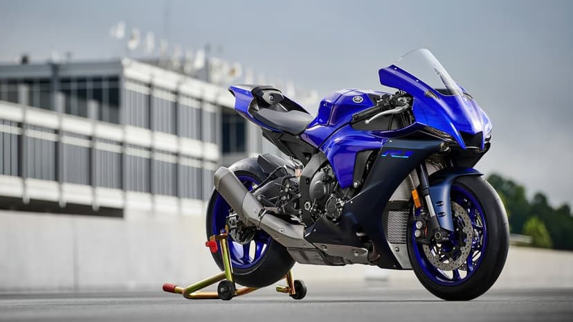 Blue Yamaha R1 motorcycle on a yellow paddock stand against a blurred racetrack building background.
