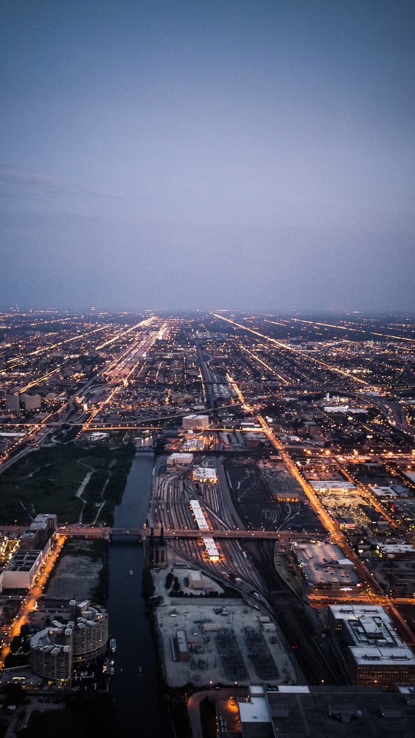 Glowing golden street grids illuminate a dark urban landscape from above near a river and railway yard at night.