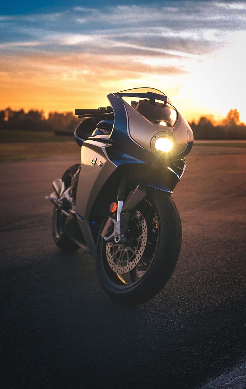 Sportbike with glowing dual headlights stands on dark asphalt under a vivid orange and purple sunset sky.