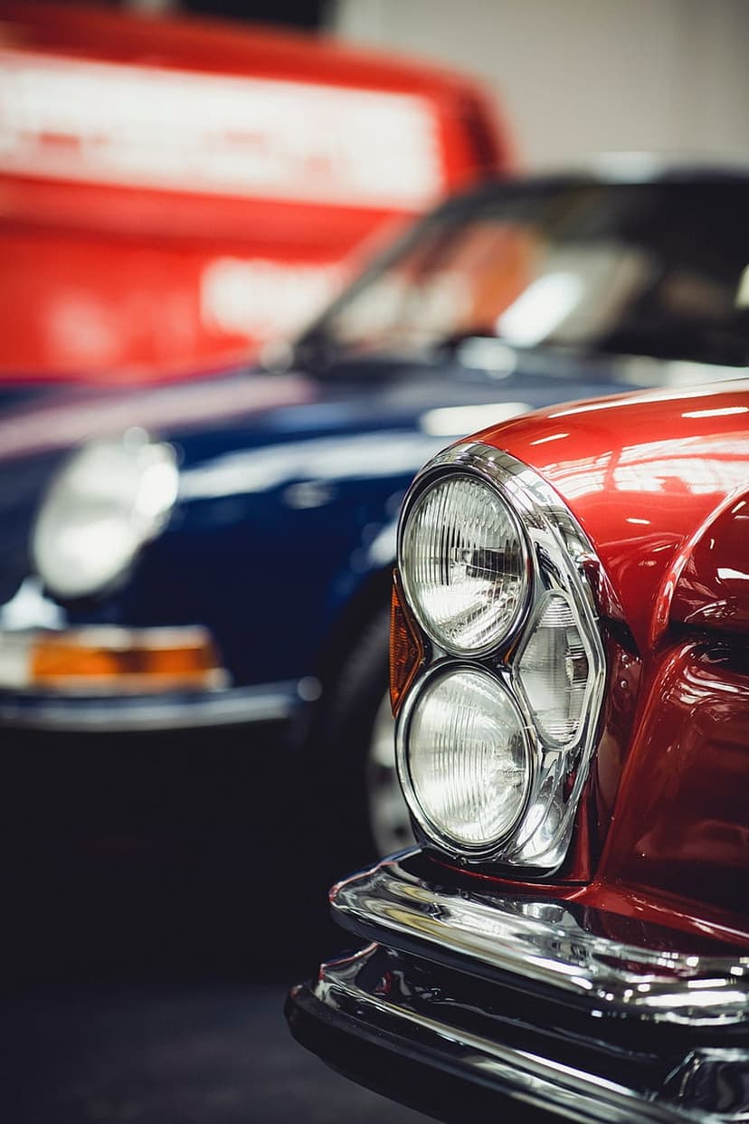 Polished chrome headlights of a red vintage car sit in front of the blurred blue body of another classic vehicle.