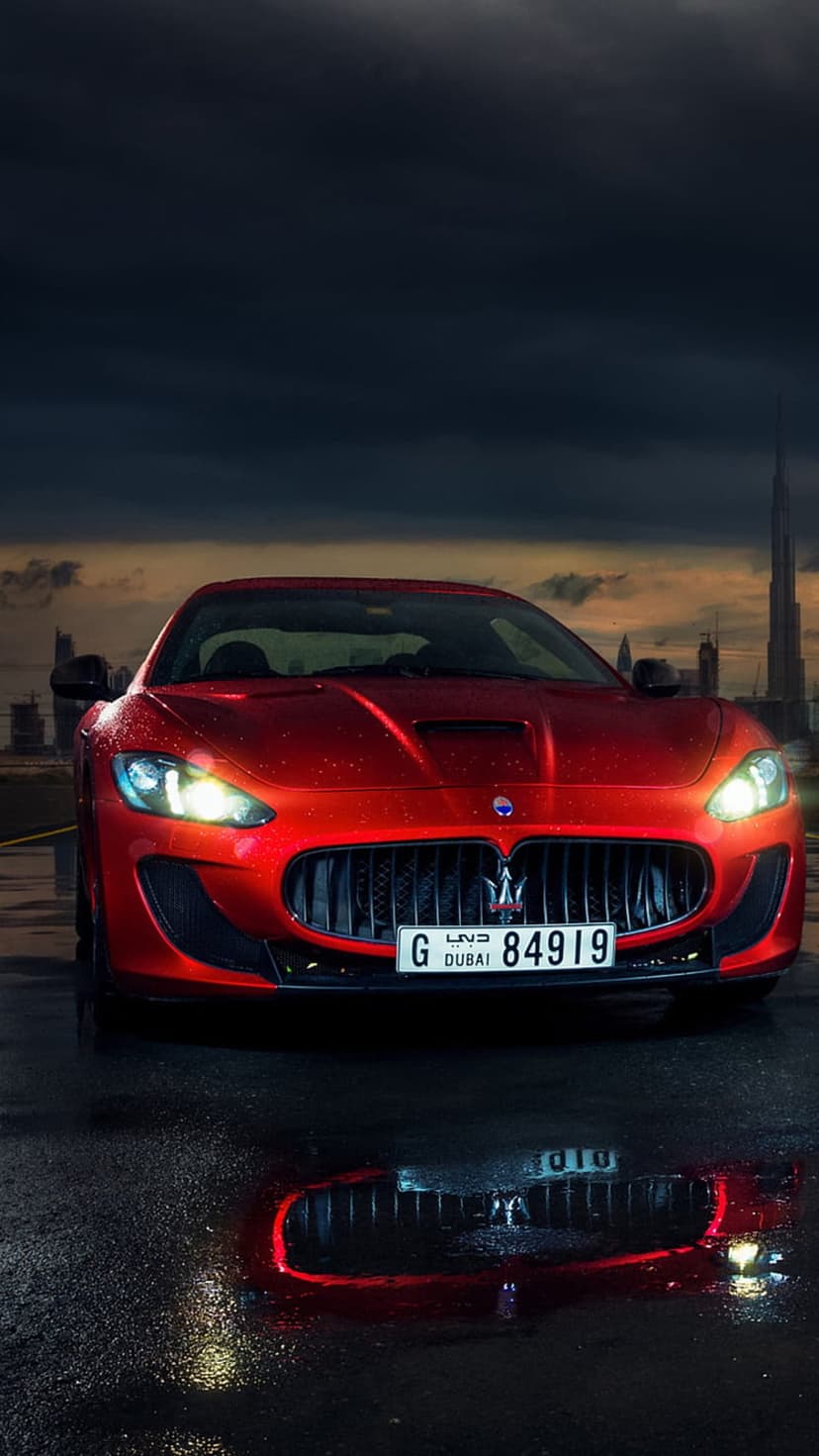 Red Maserati GranTurismo on wet Dubai asphalt at night with Burj Khalifa reflections and glowing headlights.