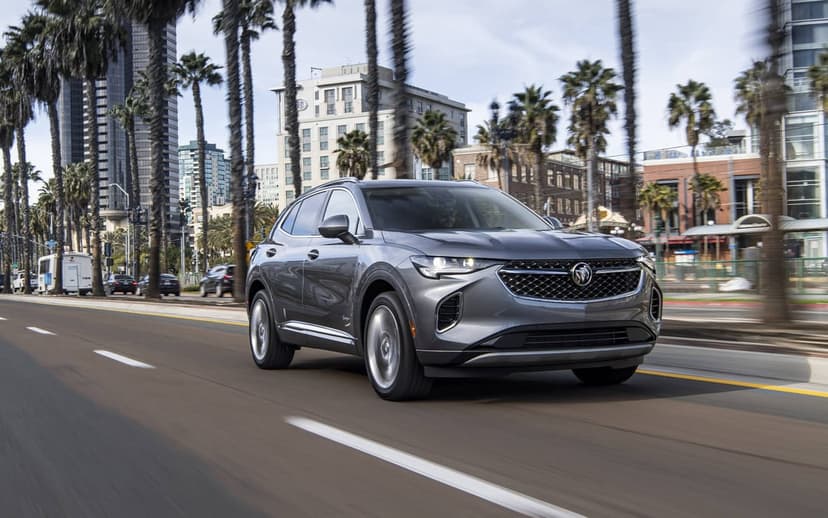 Metallic grey Buick SUV driving on an urban road lined with tall palm trees and modern high-rise buildings.