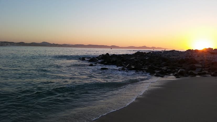 Radiant sunset illuminating breaking ocean waves, jagged coastal rocks, and hazy distant mountain peaks.