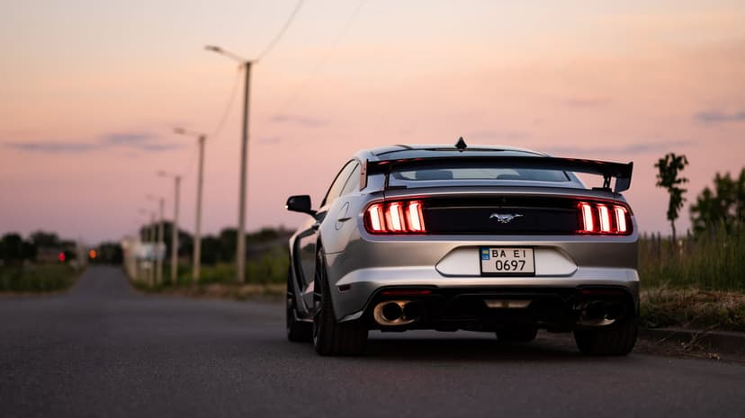 Silver Ford Mustang GT rear view with glowing taillights parked on a quiet road during a vibrant pink sunset.