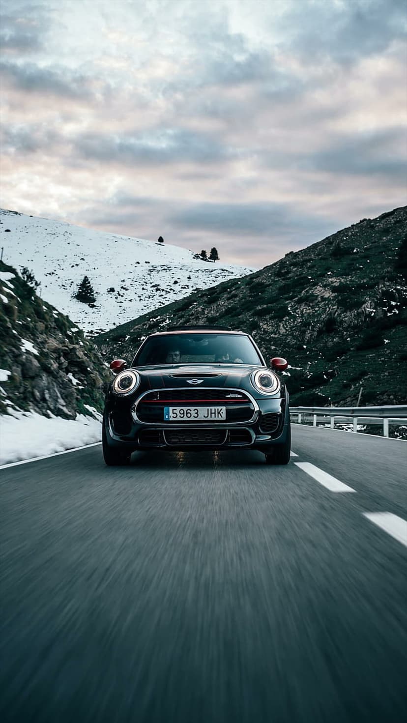 Black Mini Cooper navigating a winding asphalt road through snowy alpine peaks under a cloudy sky at dusk.