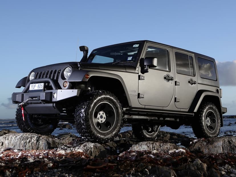 Matte charcoal Jeep Wrangler with off-road tires and snorkel parked on jagged rocks by the blue sea.