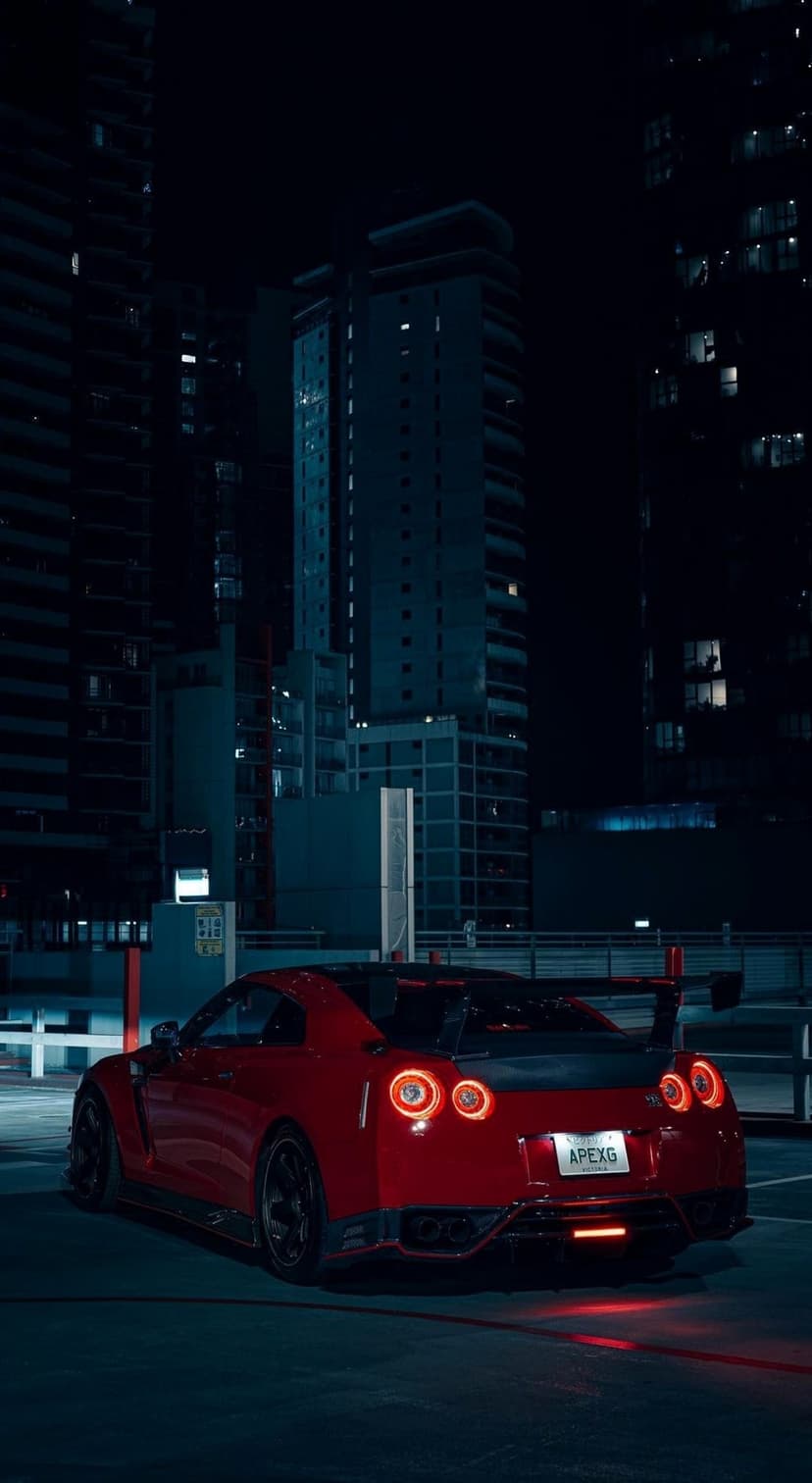Red Nissan GT-R parked on wet asphalt with glowing taillights and dark city buildings in the background.