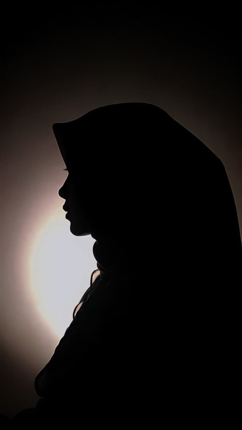 Muslim woman in a hijab stands in profile against a circular bright light, creating a dark silhouette.