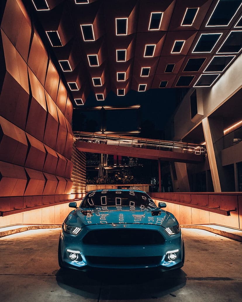 Teal Ford Mustang parked under a geometric ceiling with bright rectangular lights at a low-angle night view.