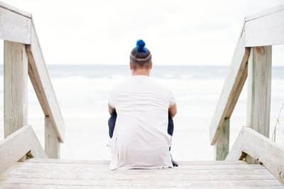 Person with blue hair sitting on beach stairs looking at ocean