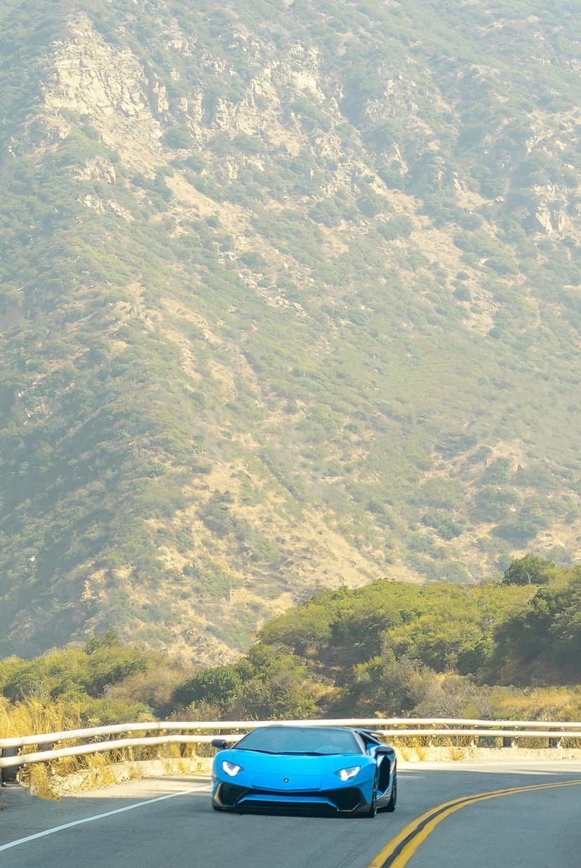 Blue Lamborghini Aventador supercar driving on a winding mountain road surrounded by green hills and sunlight.