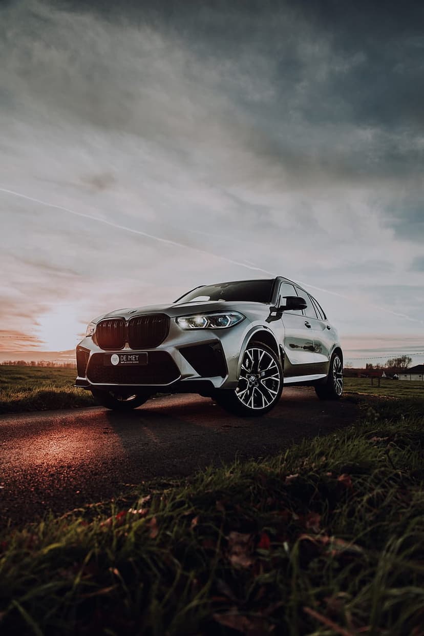 Silver BMW SUV parked on a winding asphalt road in a grassy field during a golden sunset with moody clouds.