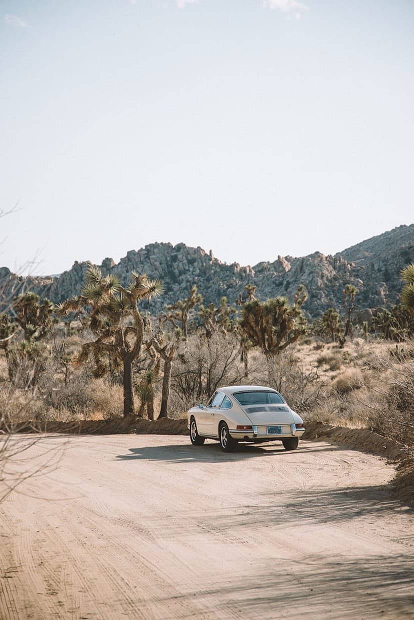 Cream Porsche 911 coupe driving on a dirt road past Joshua trees and mountains under a clear blue desert sky.