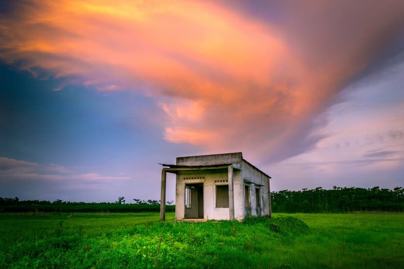 Solitary weathered house in a green field under a vast sky of glowing orange and pink sunset clouds.