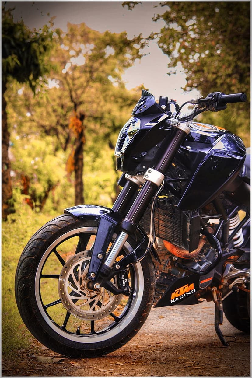 KTM Duke motorcycle in dark blue parked on a dirt trail with sunlight filtering through lush green forest trees.