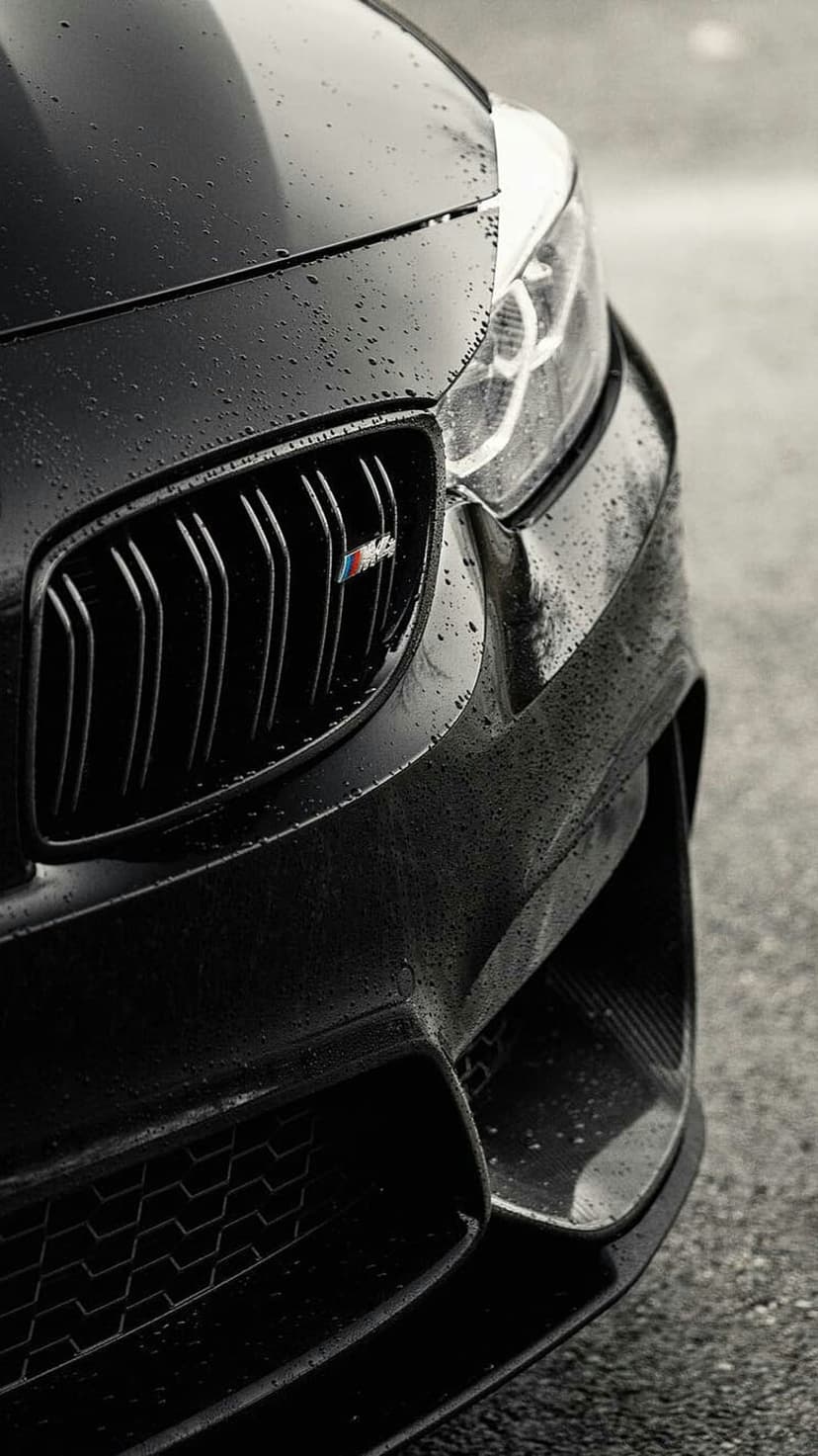 Black BMW M car headlight and front grille covered in raindrops on a wet road at night with moody lighting.