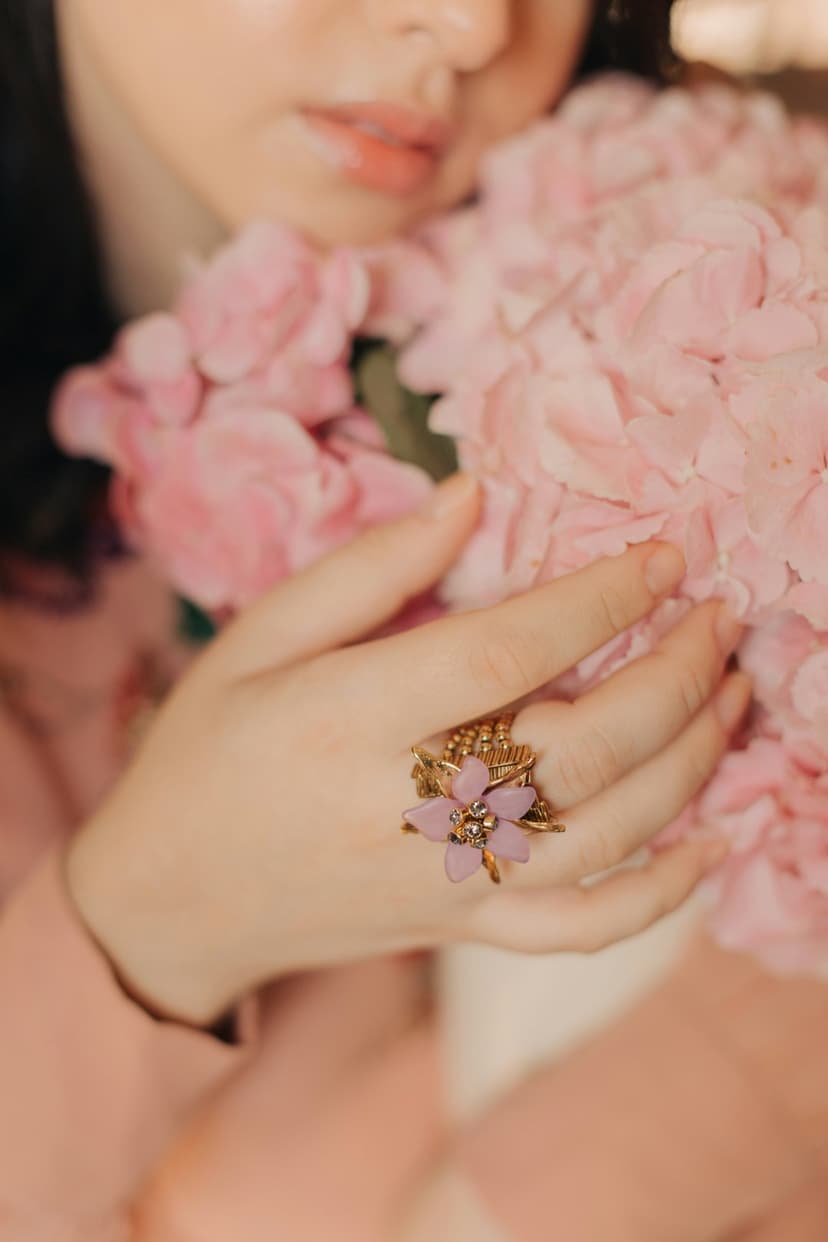 Woman's hand wearing a gold pink flower ring gently holding light pastel hydrangeas in soft natural lighting.
