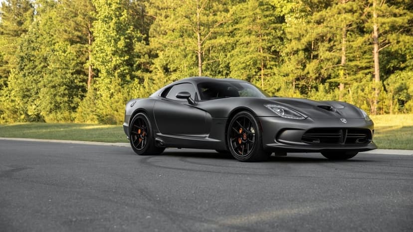 Matte black Dodge Viper GTS with orange brake calipers on a road against a lush green forest background.