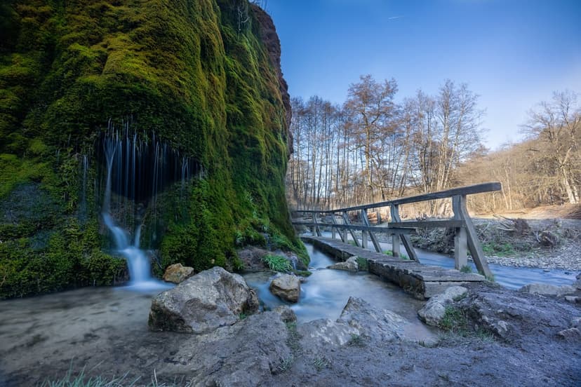 Mossy cliff waterfall and a weathered wooden footbridge over a clear forest stream under a bright blue sky.