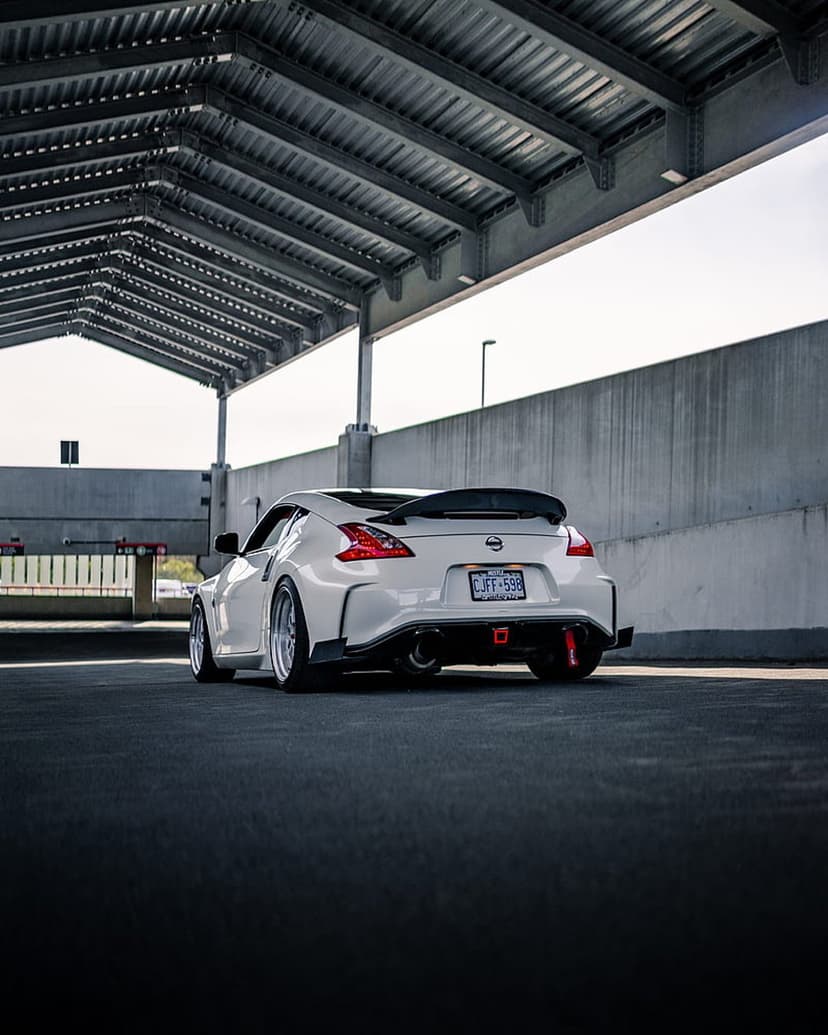 White Nissan 370Z with a rear spoiler parked under a concrete industrial shelter featuring custom alloy wheels.